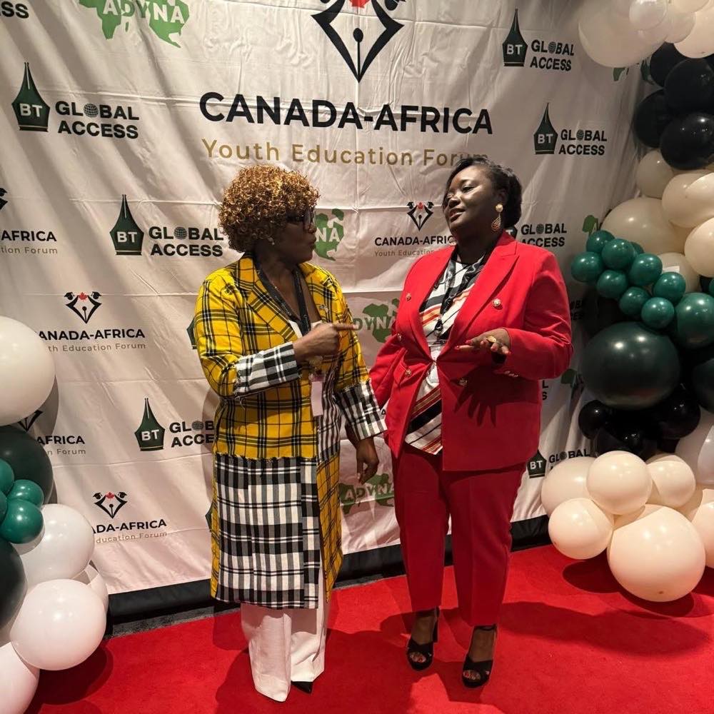 Two delegates in conversation on the red carpet in front of the forum backdrop, one gesturing mid-sentence.