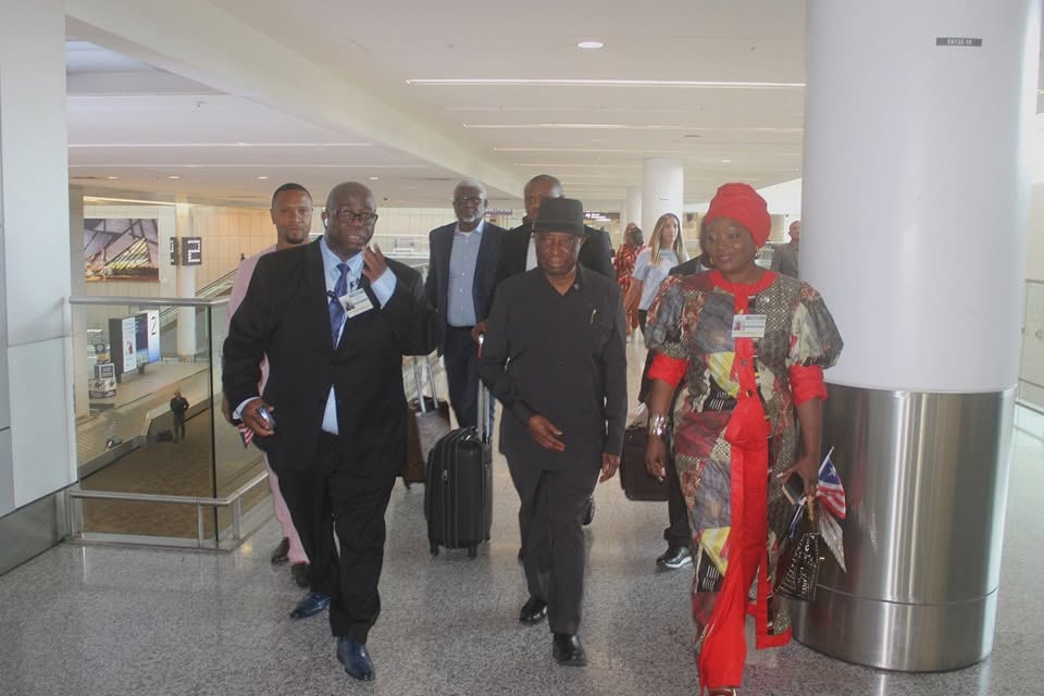 President Boakai, in a dark hat and suit, walks through an airport terminal at the centre of a group of officials wearing lanyards, with a woman in a red head wrap and patterned dress carrying a small Liberian flag beside him and wheeled luggage behind.