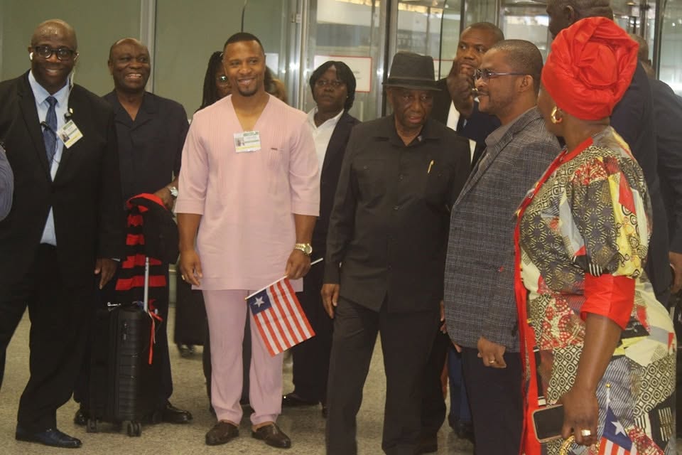 The delegation stands together inside the terminal. A man in a rose-coloured traditional suit holds a small Liberian flag on the left, President Boakai stands at the centre in a black hat, and suitcases and coats are gathered around them.