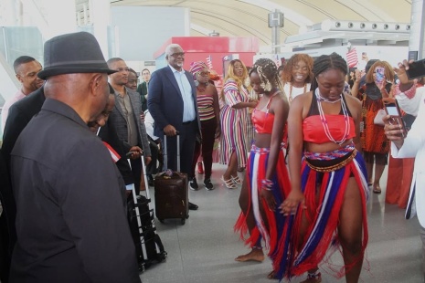 Seen from behind, President Boakai in a black hat watches young dancers in red, white and blue costumes perform in the arrivals hall while onlookers film on their phones.