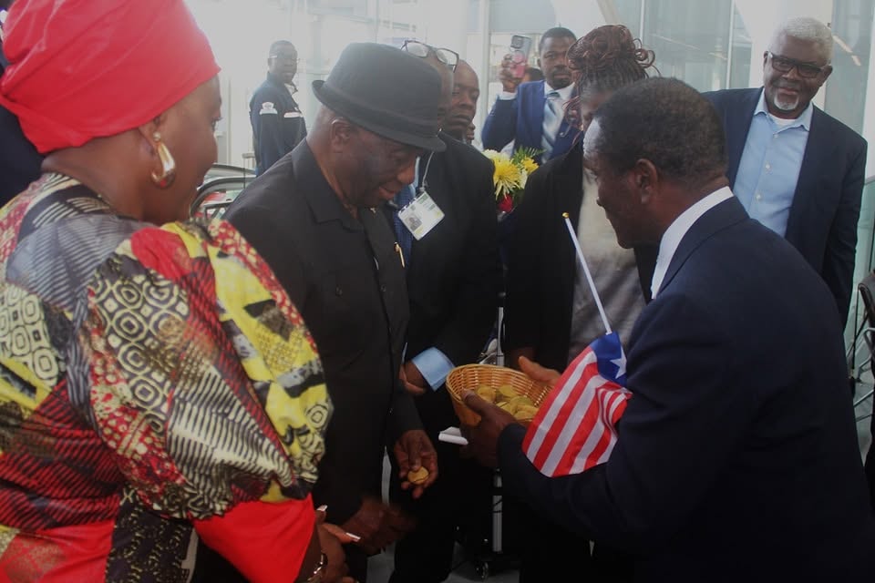A man in a dark suit holds out a small woven basket of kola nuts and a Liberian flag to President Boakai, who reaches towards it. A woman in a red head wrap and boldly patterned dress stands in the foreground and a bunch of flowers is visible behind.