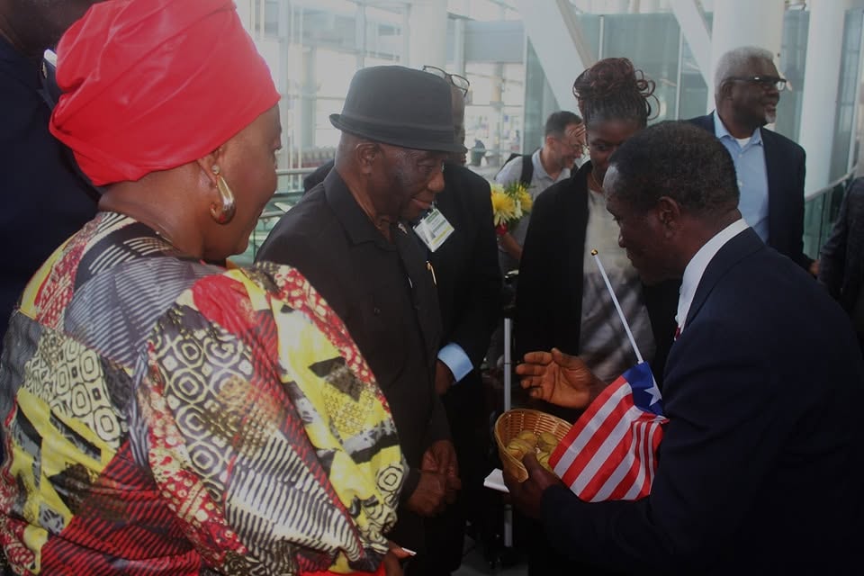 The kola nut welcome from a second angle. A woman in a red head wrap and a yellow, red and black patterned dress fills the foreground while President Boakai takes a nut from the basket held towards him.