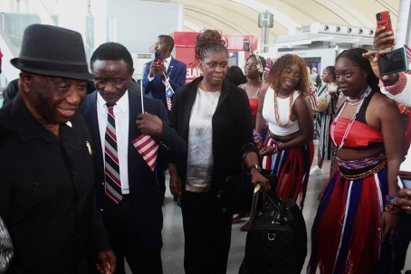 President Boakai in a black hat walks at the left of frame alongside members of the welcoming party, among them women in dark coats and dancers wearing the national colours, while a woman at the right records the moment on a phone.
