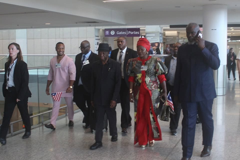 The delegation walks beneath a purple overhead sign reading Gates and Portes. President Boakai is at the centre in a black hat, a woman in a red head wrap and long red dress walks beside him, and a man in a dark suit talks on a phone at the right.