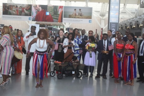 A wide view of the welcoming crowd in the arrivals hall: supporters in red, white and blue, dancers in flag-coloured skirts, someone holding flowers and a child in a pushchair, with large backlit photographic panels on the wall behind.