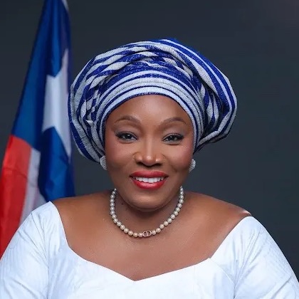 Hon. Jallahqueena Hawa Konneh, in a white dress with blue and white striped trim and a matching headwrap, before the flag of Liberia.